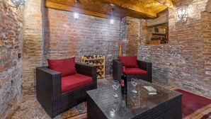 Une pièce chaleureuse aux murs de briques, meublée de meubles en osier agrémentés de coussins rouges, et dotée d’une table en verre sur laquelle reposent des verres à vin et une bouteille.