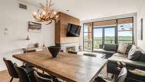 Un salon moderne avec une table à manger en bois, des chaises noires, un canapé, une cheminée et une grande fenêtre donnant sur l’extérieur.
