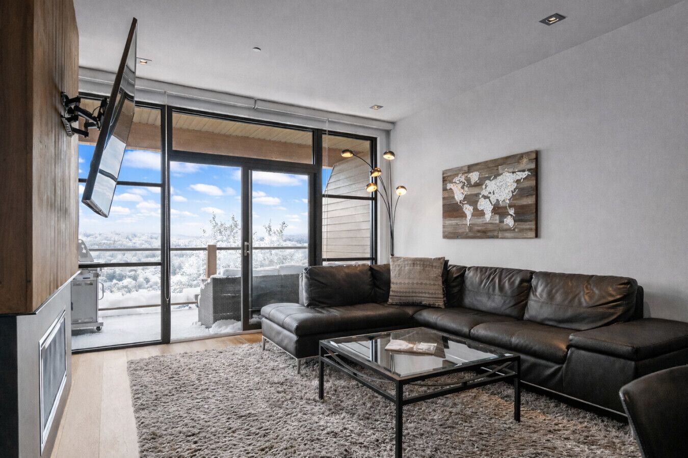A modern living room with a leather sofa, a glass coffee table, and a large sliding glass door leading to a snowy outdoor area.