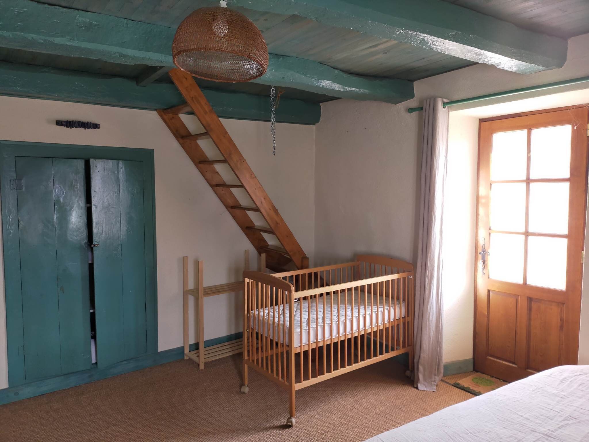Een kamer met een houten ledikant, een groene deur, een raam met gordijnen en een ladder naar een hoger niveau.