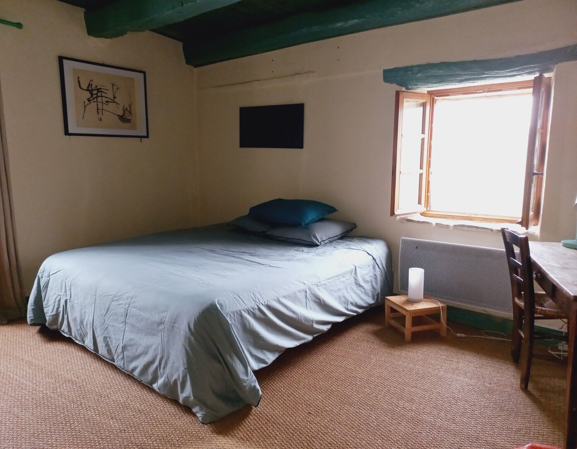 Een kamer met een bed, een klein tafeltje, een stoel, een raam en een ingelijste foto aan de muur.