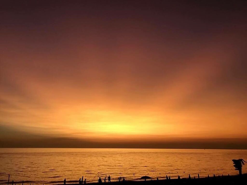 Ein Sonnenuntergang über einem ruhigen Meer mit Silhouetten von Personen und einem Boot am Ufer.
