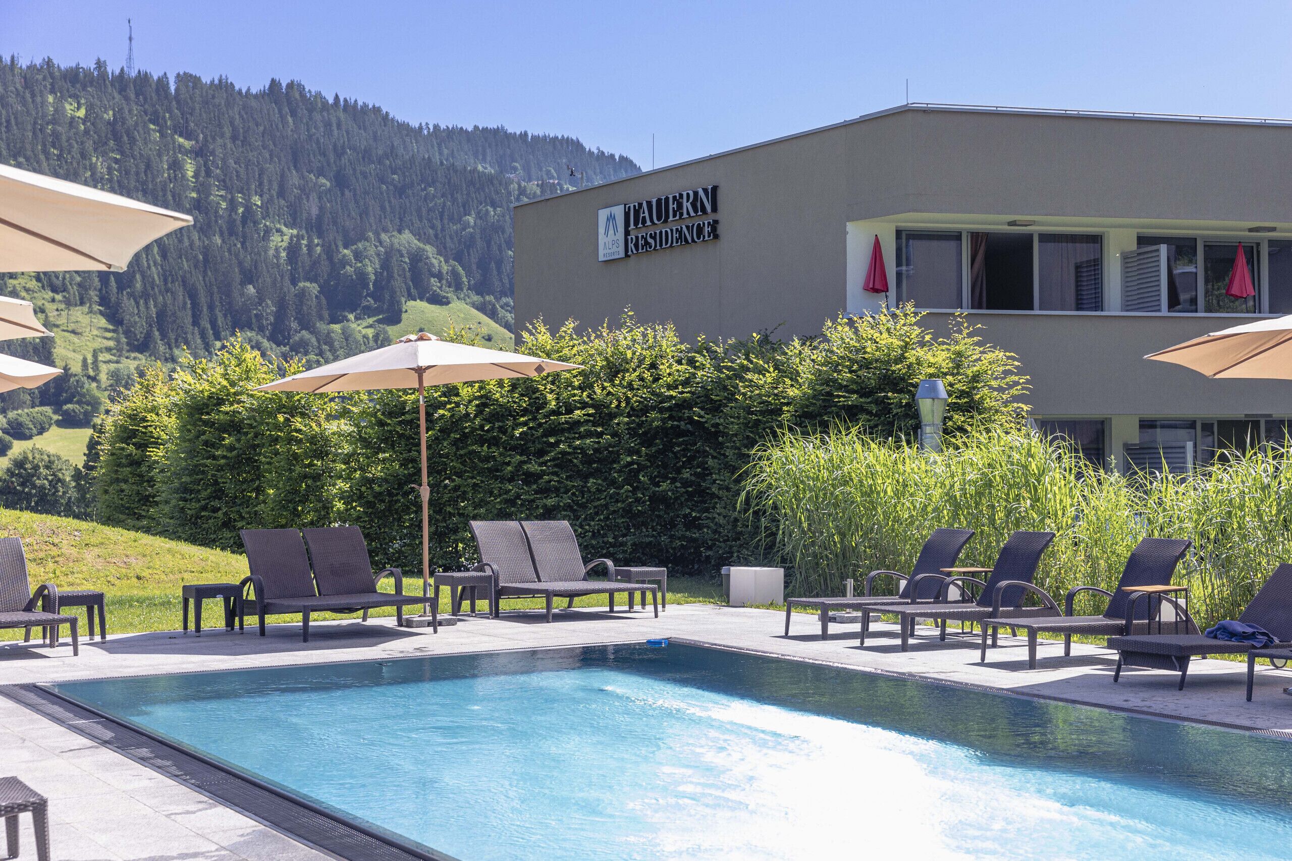 A swimming pool with lounge chairs and umbrellas, surrounded by greenery and a building with a sign reading "TAUERN RESIDENCE".