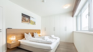 A modern bedroom with a wooden headboard, a large bed with white bedding, a bedside table with a lamp, and a window with blinds. - Superior apartment with 1 bedroom & summer pool (Radstadt)