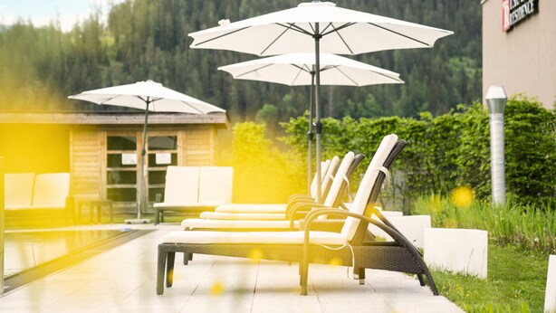 A poolside area with lounge chairs and umbrellas, a wooden building, and a forested backdrop. - Superior apartment with 2 bedrooms & summer pool (Radstadt)