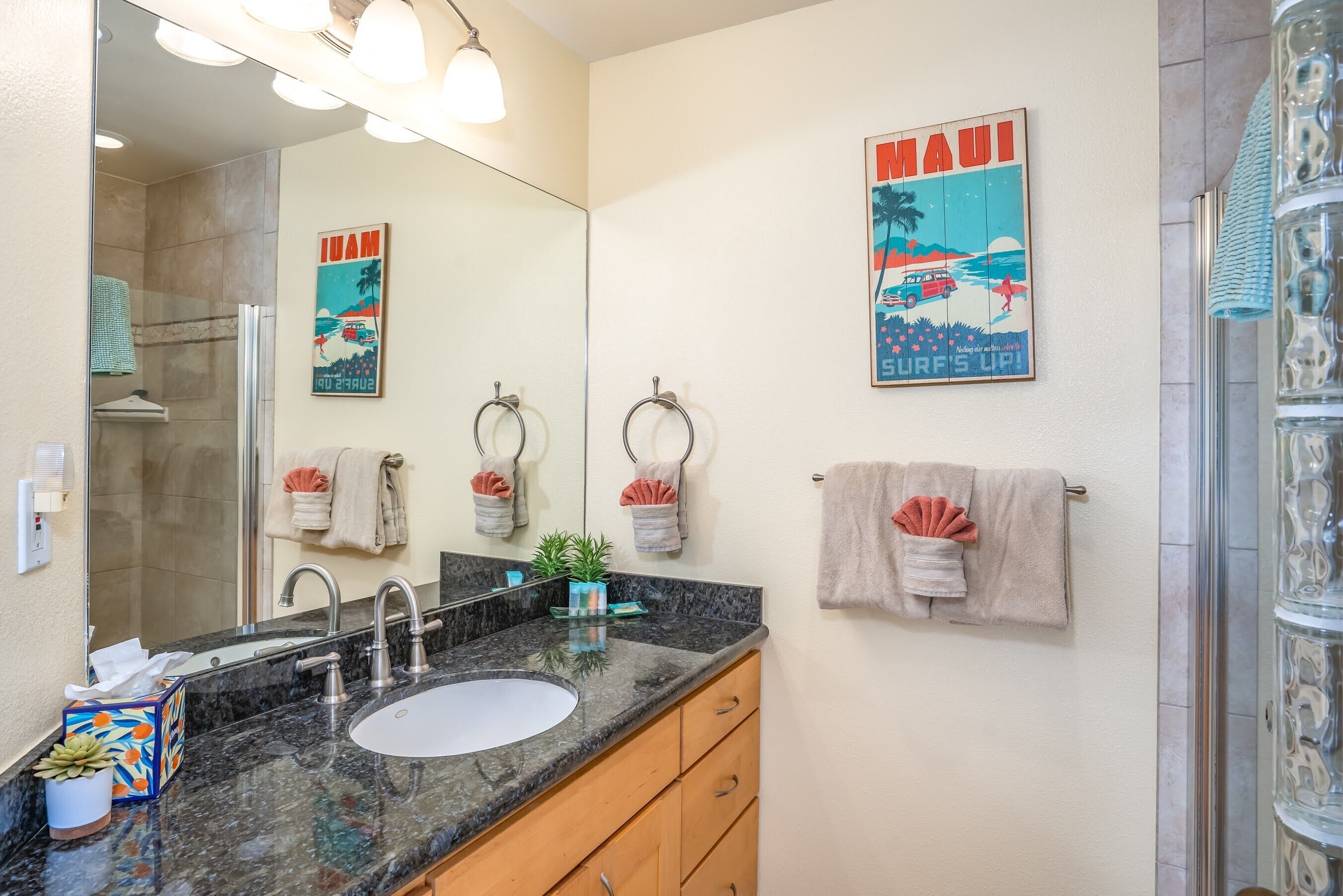 Un baño con un espejo grande, un lavabo y una ducha. Hay toallas colgadas en la pared y un cartel con una escena tropical.