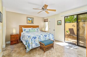 A bedroom with a bed, a nightstand, a chair, a bench, a ceiling fan, and a sliding glass door leading to an outdoor area.