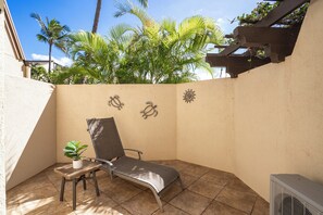 A sunlit patio with a lounge chair, a small table with a plant, and decorative wall art. - Maui Kamaole #J-119 Oceanview, Ground Flr, Great Location, near beach, A/C (Kihei)