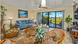 A spacious living room with a blue sofa, wicker chairs, a coffee table, and a large window offering a view of the outdoors. - Maui Kamaole #J-119 Oceanview, Ground Flr, Great Location, near beach, A/C (Kihei)