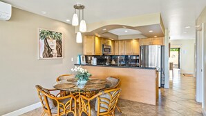 A modern kitchen with wooden cabinets, stainless steel appliances, and a dining area with wicker chairs and a glass table. - Maui Kamaole #J-119 Oceanview, Ground Flr, Great Location, near beach, A/C (Kihei)