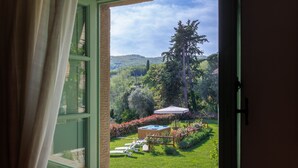 Vista da una finestra con tende su un giardino rigoglioso, con idromassaggio, terrazza in legno e montagne sullo sfondo.
