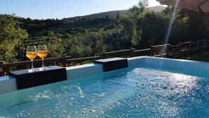 Una piscina con due bicchieri di vino sul bordo, circondata da un pontile in legno e affacciata su un paesaggio rigoglioso.