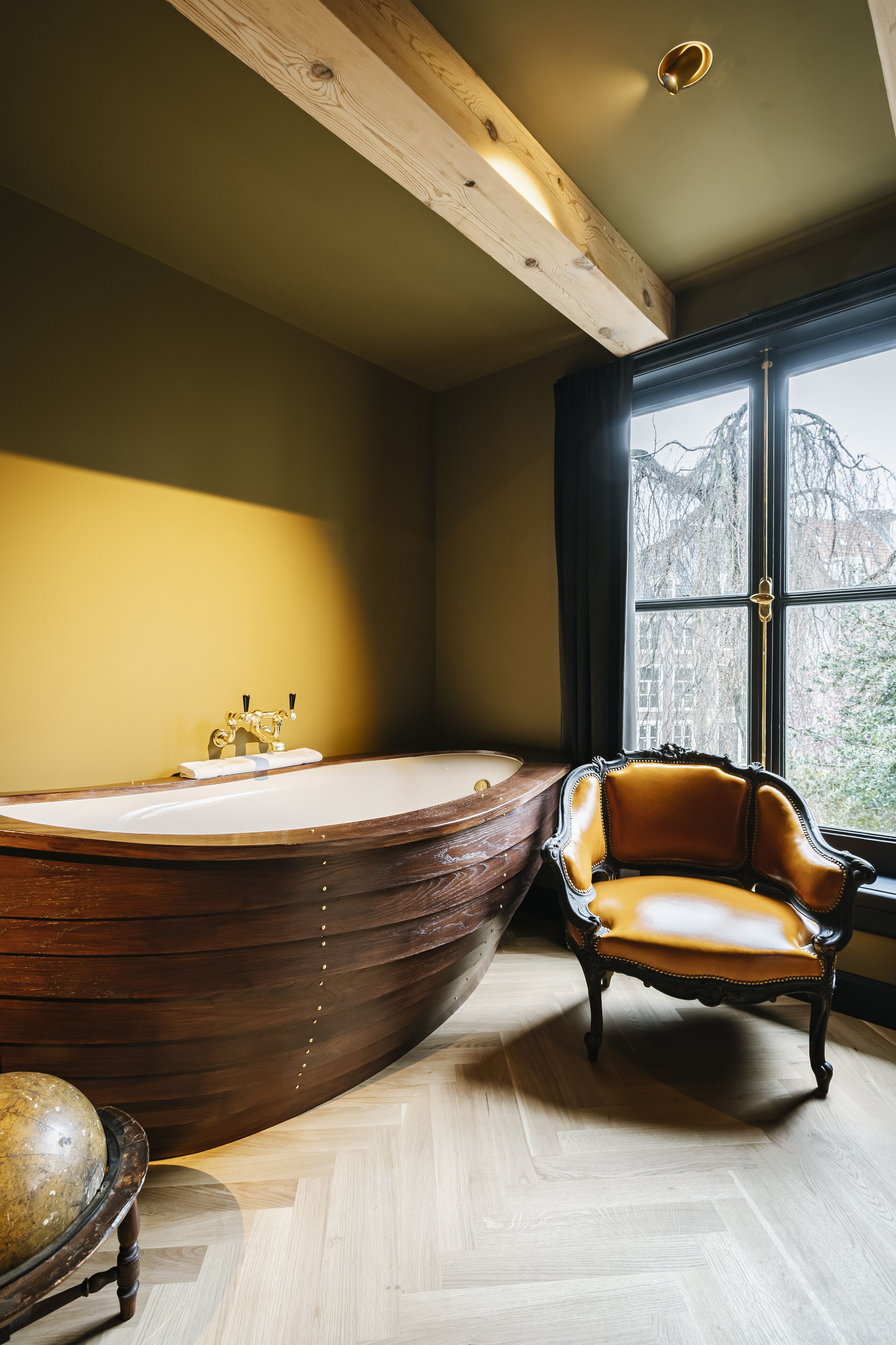 A bathroom with a wooden boat-shaped bathtub, a classic armchair, and a large window with a view of trees.