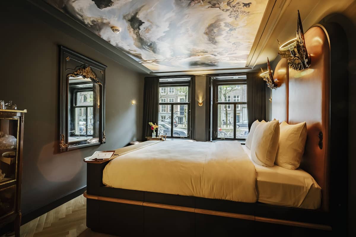 A bedroom with a large bed, a ceiling mural, and two windows with views of buildings.