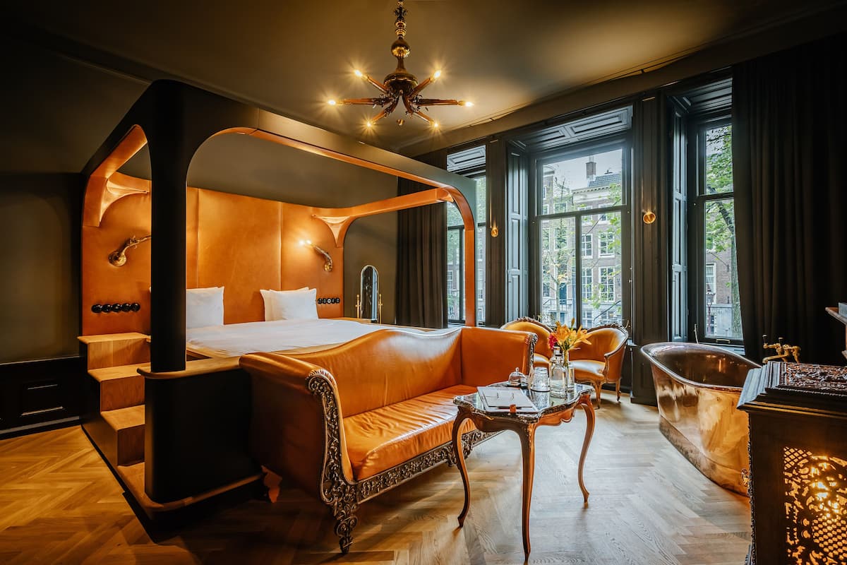 A luxurious bedroom with a four-poster bed, a sitting area with a sofa, a wooden coffee table, and a vintage bathtub.