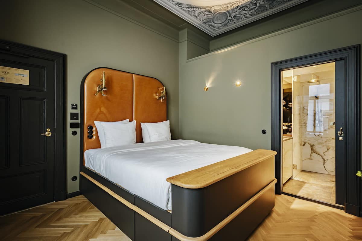 A modern bedroom with a wooden headboard, a large bed, a wooden bedside table, and a marble bathroom visible through an open door.