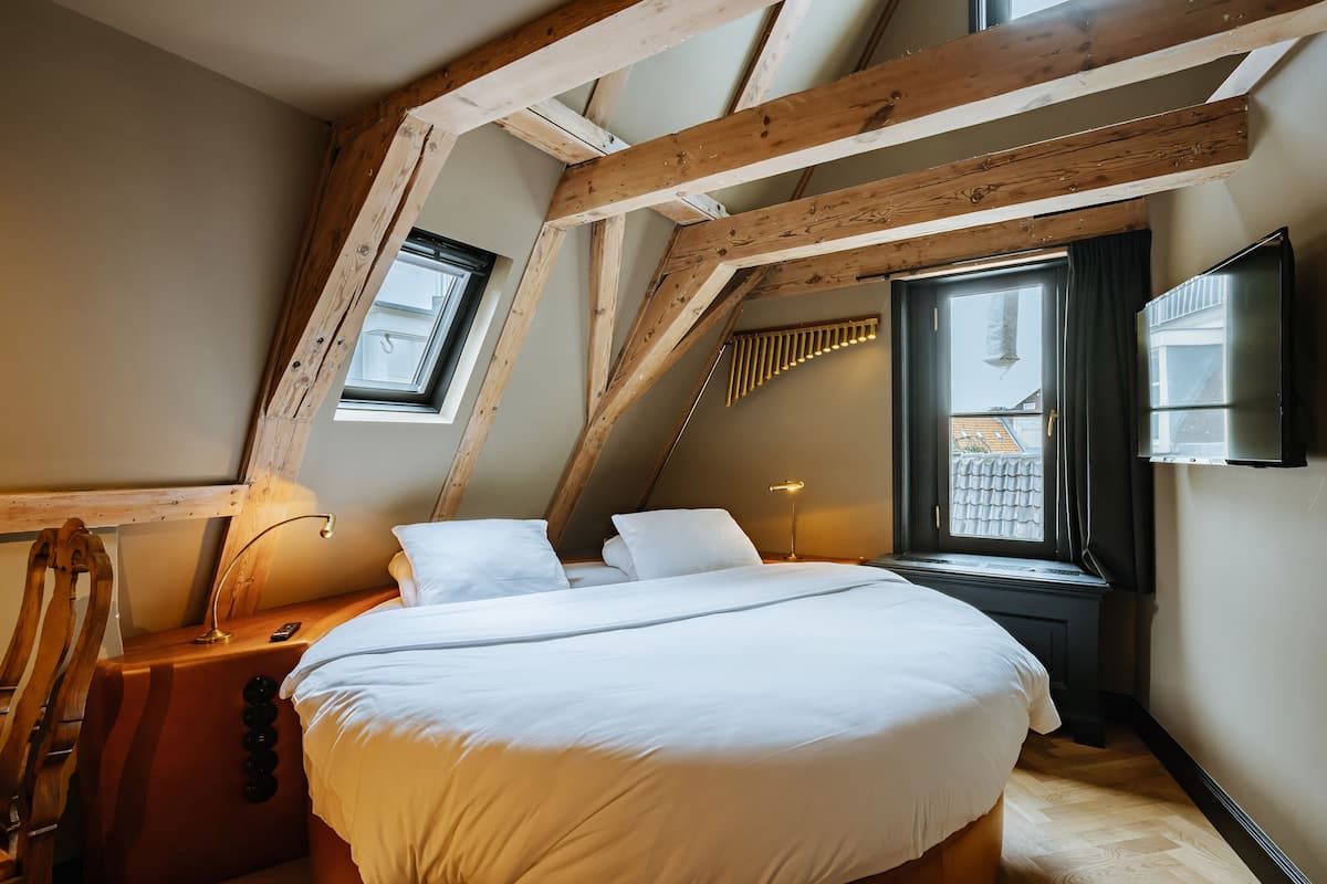 A bedroom with a sloped ceiling, wooden beams, a bed with white bedding, a nightstand, a lamp, and a window with curtains.