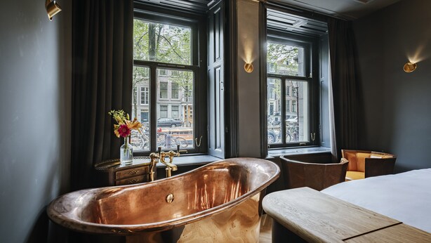 A modern bathroom with a large copper bathtub, a wooden bench, and a view of the city through large windows. - Hotel The Noblemen (Amsterdam)
