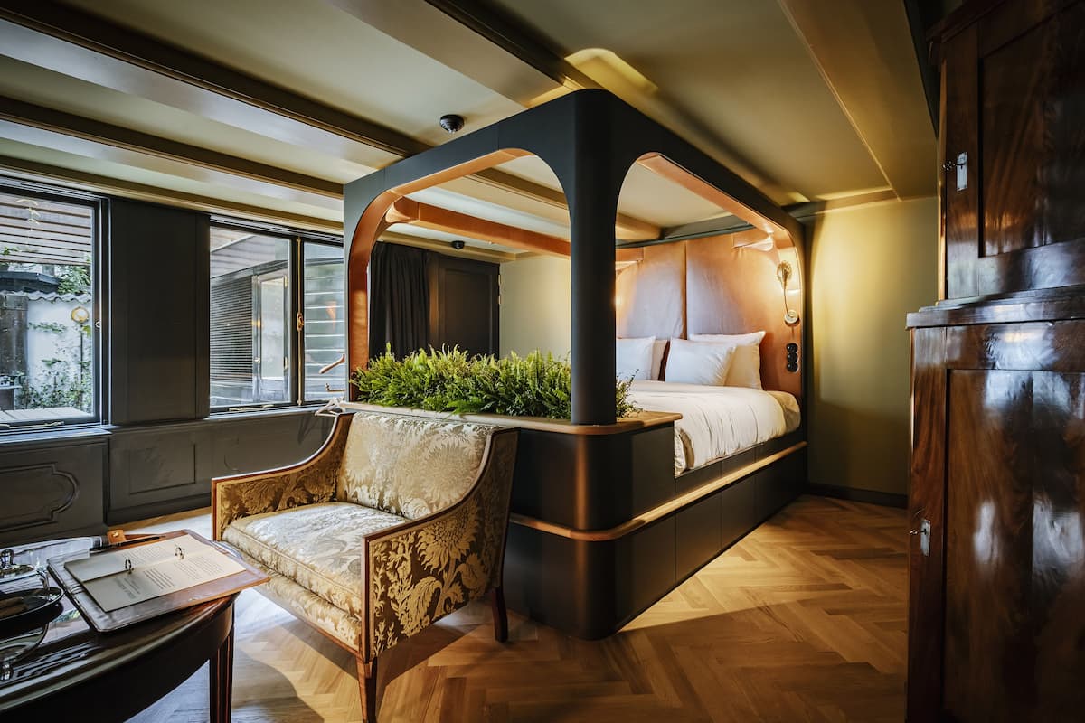 A modern bedroom with a canopy bed, a seating area with a chair, and a desk with a clipboard.