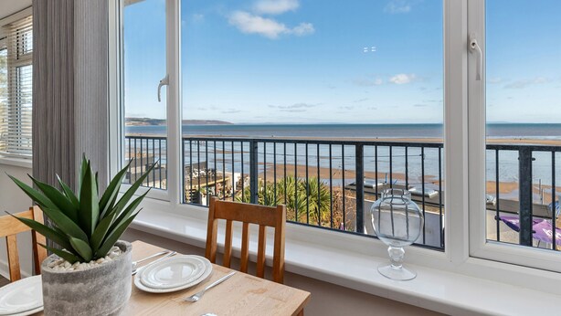 Un balcón con vista a la playa, una mesa de comedor con platos y una copa, y una planta en maceta.