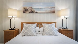 A neatly made bed with two bedside lamps, a wooden headboard, and a painting of a beach scene above it. - Sea View Apartment - Sea Front Apartment With Views (Saundersfoot)