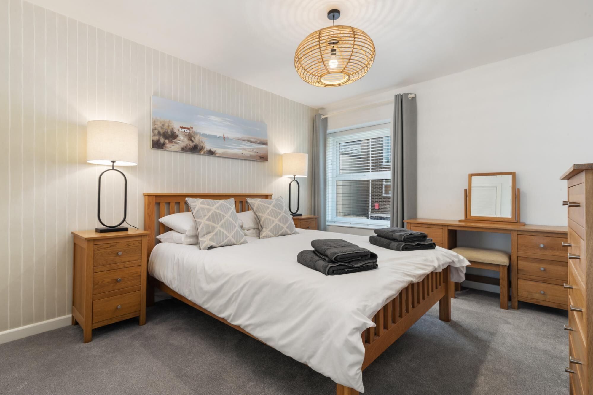 Une chambre à coucher bien rangée, avec un lit en bois, des tables de chevet, une commode et une grande fenêtre équipée de stores.