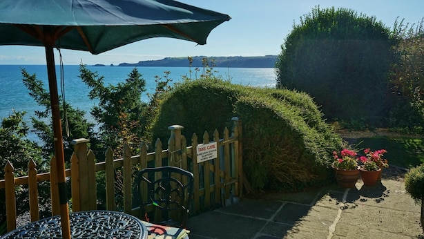 En uteplats med ett bord och stolar, ett trästaket, en skylt som uppmanar till försiktighet på hala trappsteg när det är vått, och utsikt över havet.