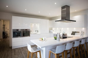 A modern kitchen with white cabinetry, a central island with bar stools, built-in ovens, and a stainless steel range hood.