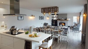A modern kitchen with a central island, dining area with white chairs, a fireplace, and a living room with a sofa and armchairs. - The Hawthorns - Large Luxury Cottage Hot Tub Sea Views (Saundersfoot)