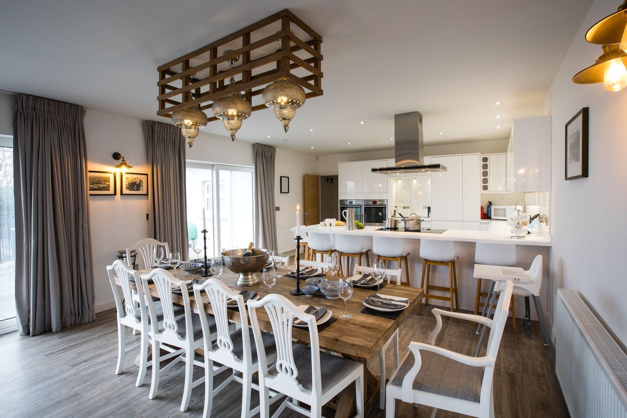 A modern dining area with a wooden table and white chairs, a kitchen in the background, and a large window with curtains.