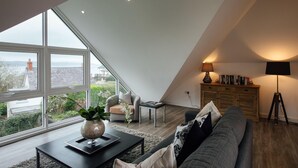 A modern loft bedroom with a large skylight, a cozy seating area with a sofa and armchair, a wooden cabinet with books, and a lamp. - The Hawthorns - Large Luxury Cottage Hot Tub Sea Views (Saundersfoot)