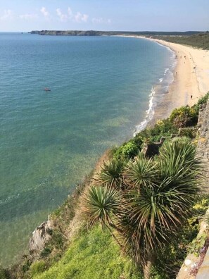 A coastal landscape with a sandy beach, clear blue waters, and green vegetation along the cliffside.
