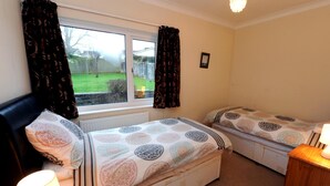 A bedroom with two beds, a window with curtains, a radiator, and a bedside lamp. - Glan Heulog - Near to Beach Pet Friendly (Saundersfoot)