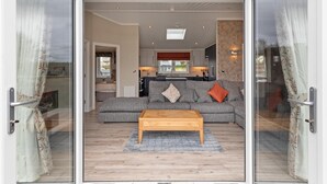 A modern living room with a grey sofa, a wooden coffee table, and a view through glass doors to a kitchen and outdoor area. - Valley View Lodge - Luxury Lodge Hot Tub Close to Beach (Tenby)