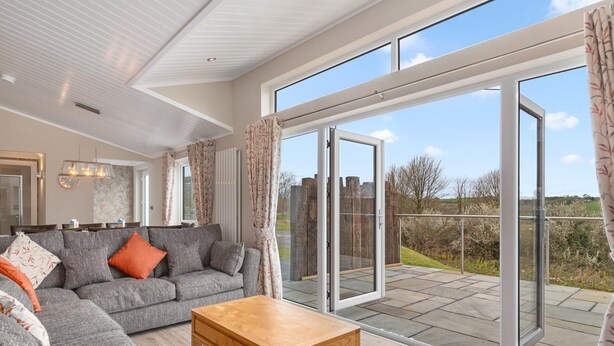 A spacious living room with a grey sofa, a wooden coffee table, and a large sliding glass door leading to an outdoor patio. - Valley View Lodge - Luxury Lodge Hot Tub Close to Beach (Tenby)