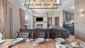A modern dining area with a large table set for breakfast, a cozy seating area with a sofa, and a kitchen in the background. - Valley View Lodge - Luxury Lodge Hot Tub Close to Beach (Tenby)