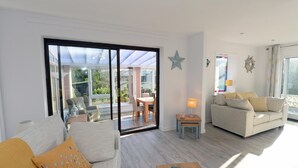 Ein modernes Wohnzimmer mit einer Couch, einem Couchtisch und einem Essbereich mit Stühlen und einem Tisch. Ein großes Schiebeglasfenster führt auf einen Balkon mit Blick auf grüne Vegetation.