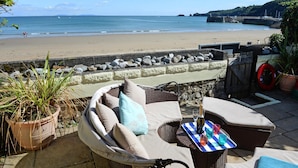 Eine Terrasse am Strand mit Korbmöbeln, einem Tisch mit Getränken und Blick auf das Meer und Boote.