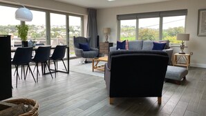 A modern living room with a skylight, grey and black furniture, a dining area with chairs, and large windows offering a view of the city. - Amber Cottage - Luxury Cottage Hot Tub Sea Views and Log Burner (Saundersfoot)