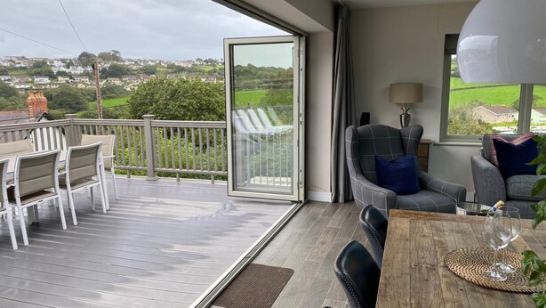 A spacious balcony with a wooden deck, a dining table with chairs, a sofa, and a large window offering a view of the countryside. - Amber Cottage - Luxury Cottage Hot Tub Sea Views and Log Burner (Saundersfoot)