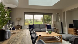 A modern living room with a skylight, a large window, a dining table, sofas, and a television. - Amber Cottage - Luxury Cottage Hot Tub Sea Views and Log Burner (Saundersfoot)