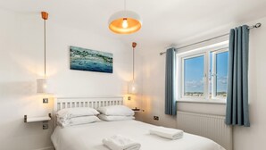 A bedroom with a bed, white bedding, a painting of a coastal scene, a window with curtains, and pendant lights. - Am Byth - Luxury Cottage Sea Views (Saundersfoot)