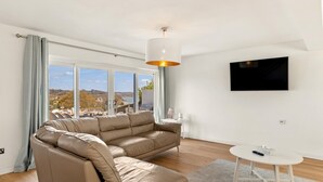 A modern living room with a beige sectional sofa, a round coffee table, and a TV mounted on the wall. - Am Byth - Luxury Cottage Sea Views (Saundersfoot)