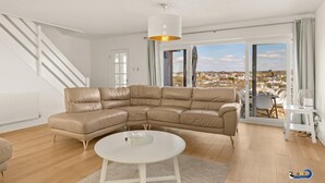 A spacious living room with a large L-shaped beige leather sofa, a white round coffee table, and a view of a town through large windows. - Am Byth - Luxury Cottage Sea Views (Saundersfoot)