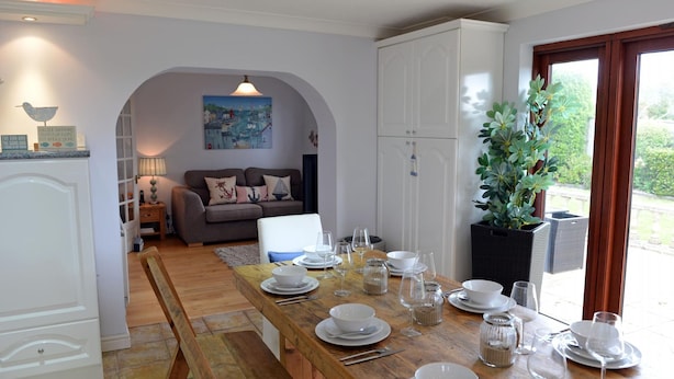 A dining area with a wooden table set for a meal, white cabinets, and a view of greenery outside. - Swallow Dale - Large Pet Friendly Cottage (Saundersfoot)