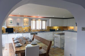 A kitchen with white cabinets, a wooden dining table set with plates and glasses, and a window with blinds. - Swallow Dale - Large Pet Friendly Cottage (Saundersfoot)