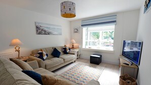 A living room with a beige sofa, two side tables with lamps, a television, a window with blinds, and a painting on the wall.