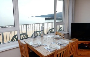 A dining table set for four with a view of the sea and a lighthouse through large windows. - Sea Urchins Apartment - Sea Front Apartment With Views Pet Friendly (Saundersfoot)