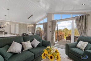 A spacious living room with a green sofa set, a dining area, and a large sliding glass door leading to an outdoor patio. - Trevelyn Lodge - Luxury Lodge Hot Tub Close to Beach (Tenby)