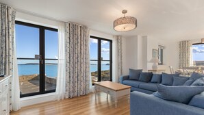 A spacious living room with a large window offering a sea view, a blue sofa, a wooden coffee table, and a hanging light fixture. - Apartment 8 Waterstone House - Luxury Apartment Sea Views Pet Friendly (Tenby)